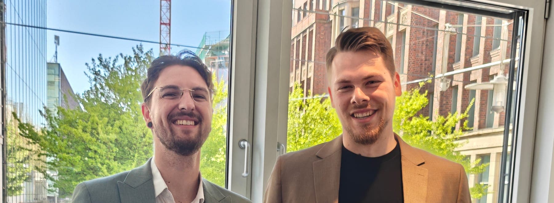 Alexander Schmiedeberg und Lukas Vogel stehen lachend nebeneinander vor einem Fenster.